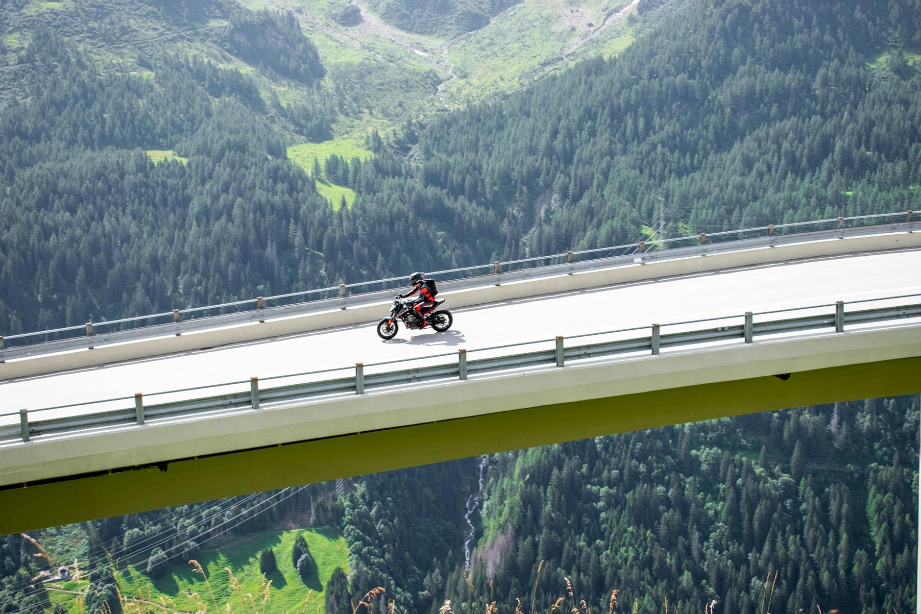 Ein Motorrad mit Fahrer in rotem Anzug fährt auf einer hohen Brücke über ein bewaldetes Gebirgstal mit Bach.