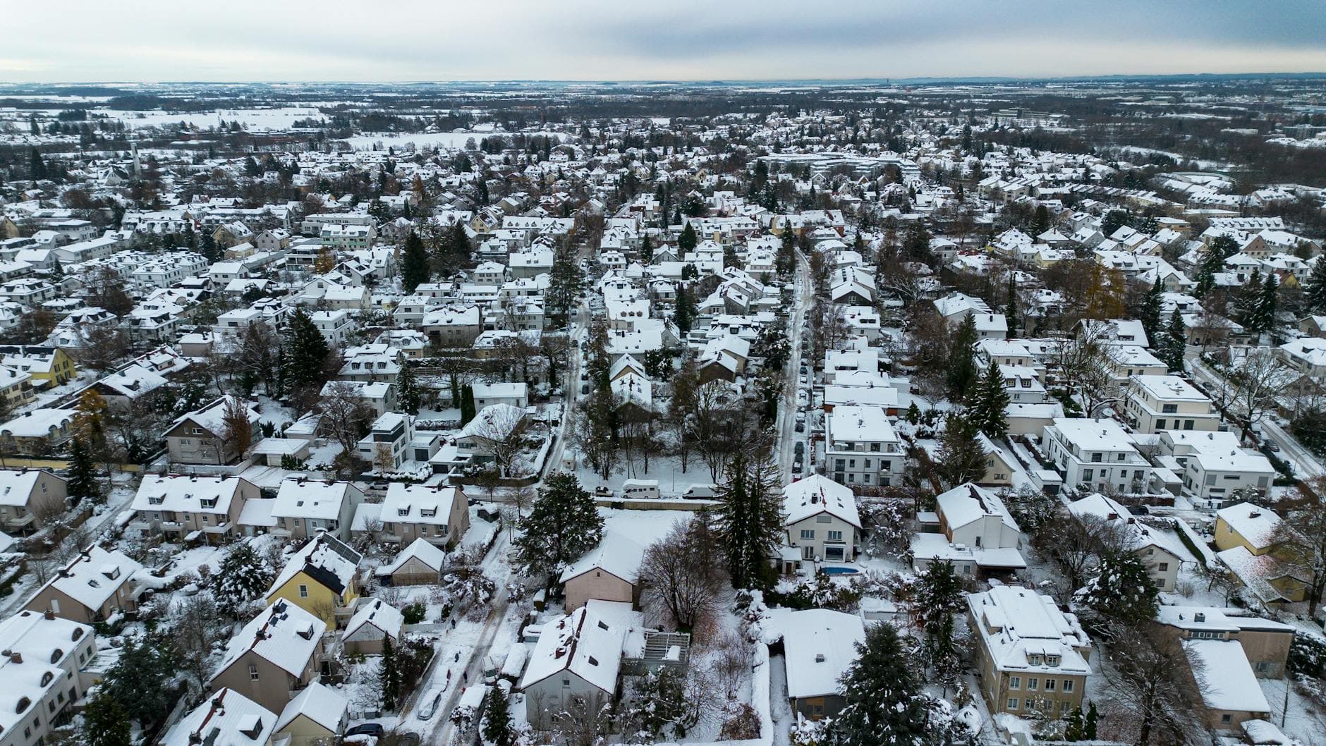 Luftaufnahme einer verschneiten Wohnsiedlung mit Häusern, Bäumen und Straßen unter bewölktem Himmel im Winter.