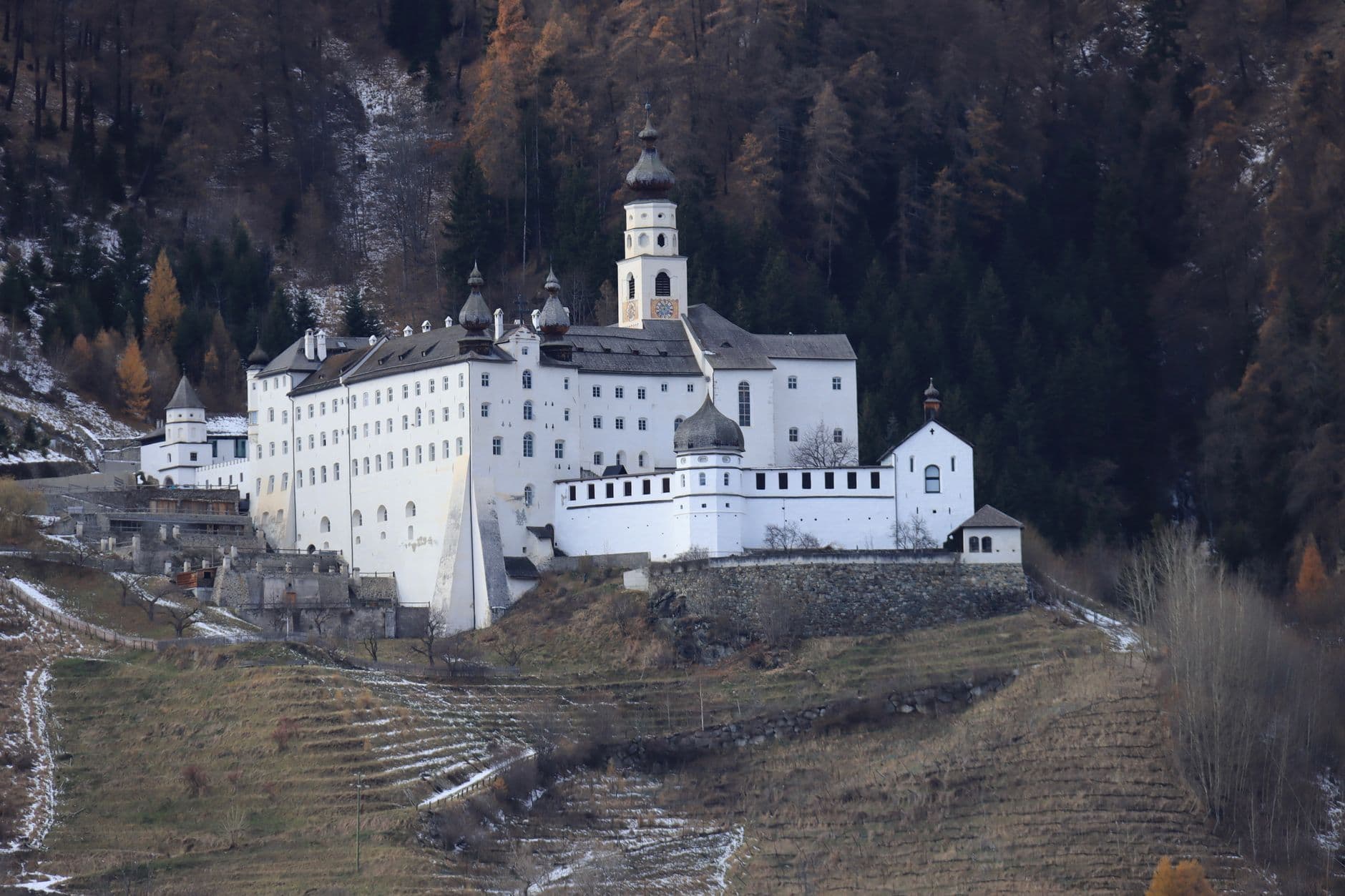 Ein großes, weißes Kloster mit Glockenturm auf einem Hügel, umgeben von terrassierten Feldern und dunklem Wald mit etwas Schnee.
