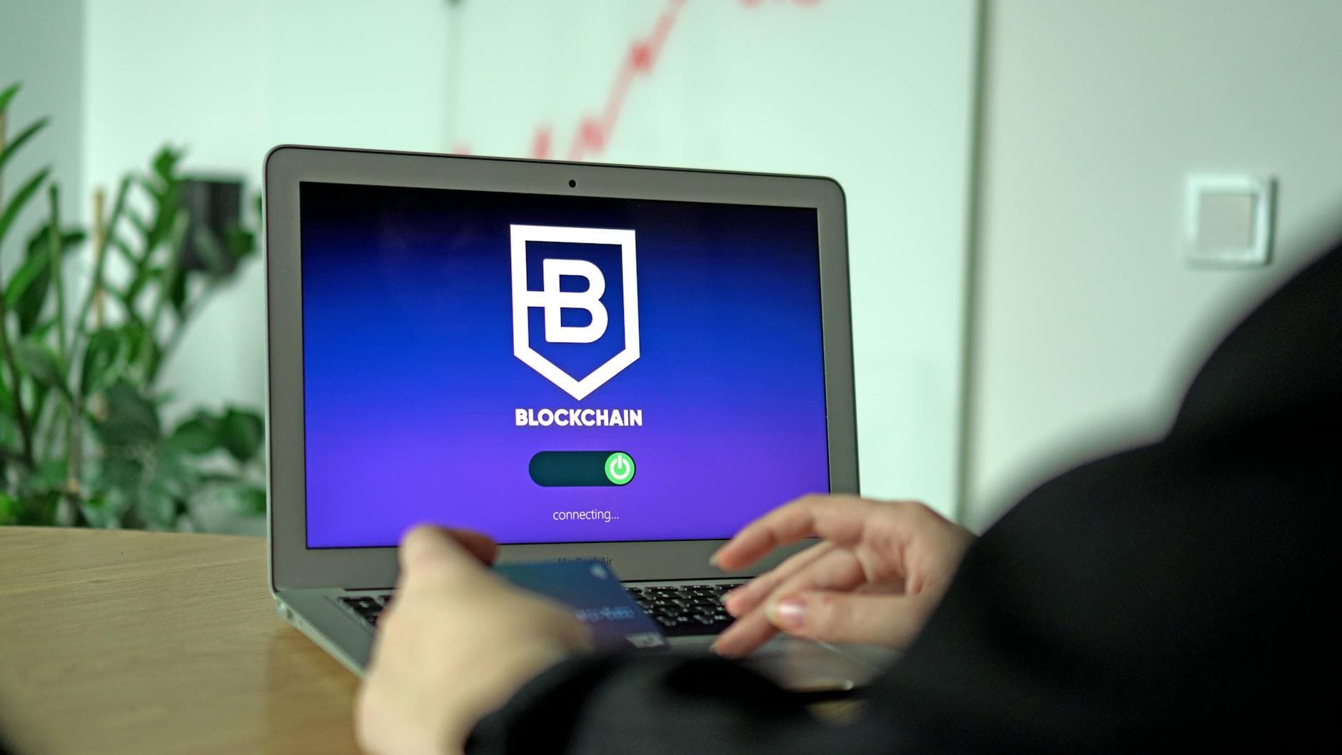Person hält Kreditkarte und bedient Laptop mit Blockchain-Logo und "connecting..."-Anzeige auf dem Bildschirm.