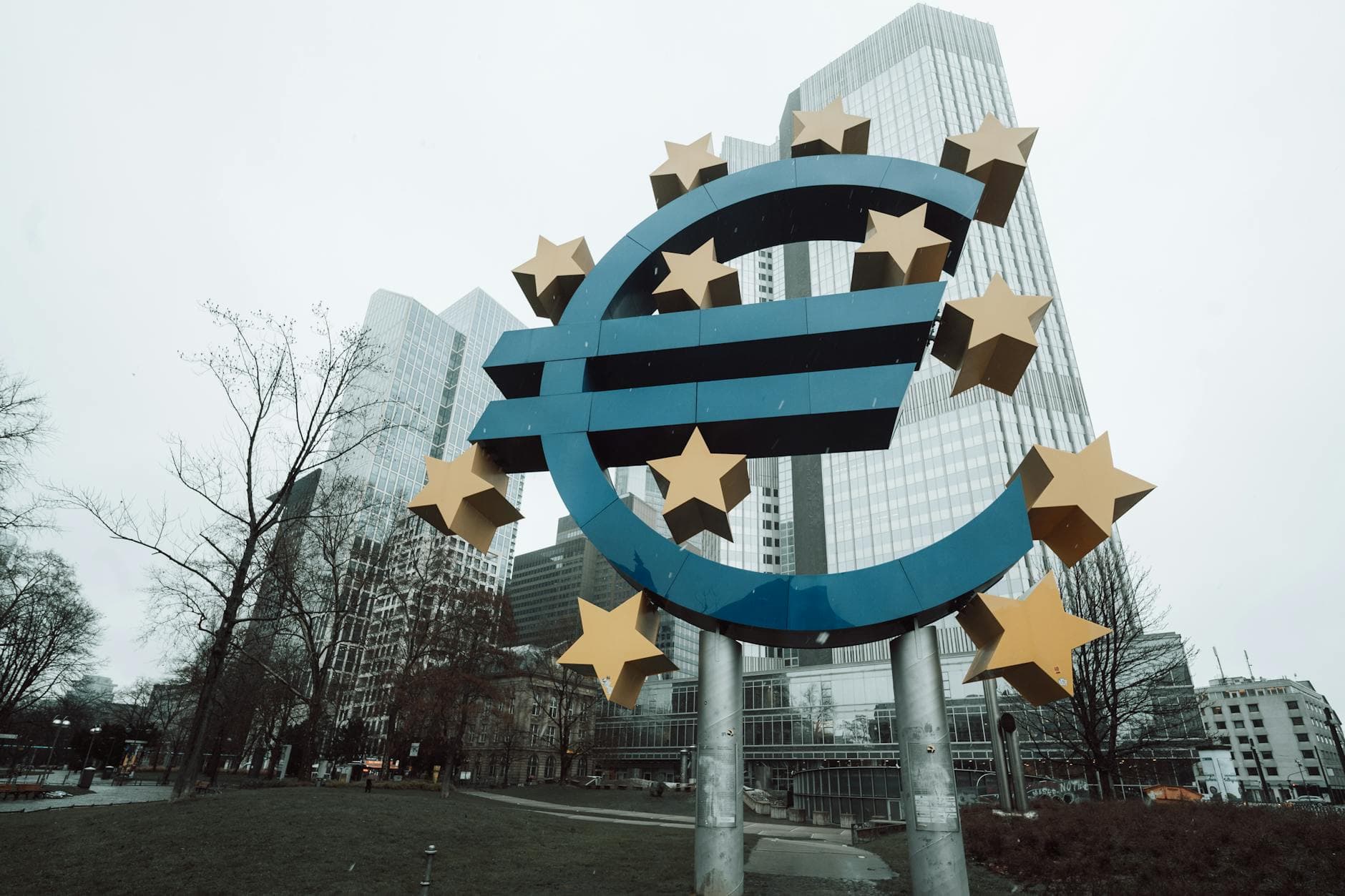 Blaue Euro-Skulptur mit goldenen Sternen vor hohen, modernen Gebäuden und kahlen Bäumen an einem trüben Wintertag.