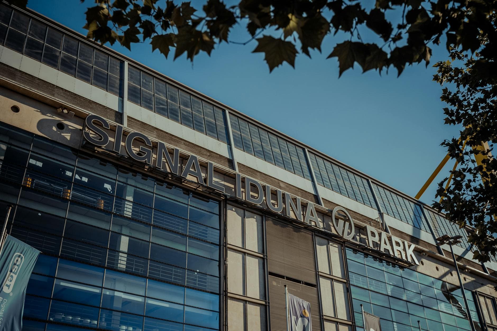 Der Schriftzug SIGNAL IDUNA PARK auf einer modernen Gebäudefassade mit Glasfenstern unter blauem Himmel und Baumblättern.