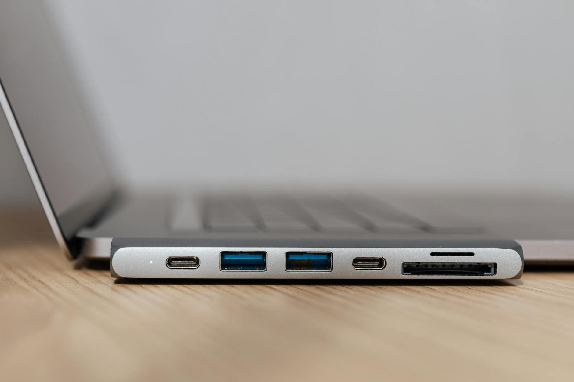 Silberner USB-C-Hub mit zwei USB-A, zwei USB-C (einer leuchtet) und zwei Kartensteckplätzen an einem Laptop auf Holztisch.