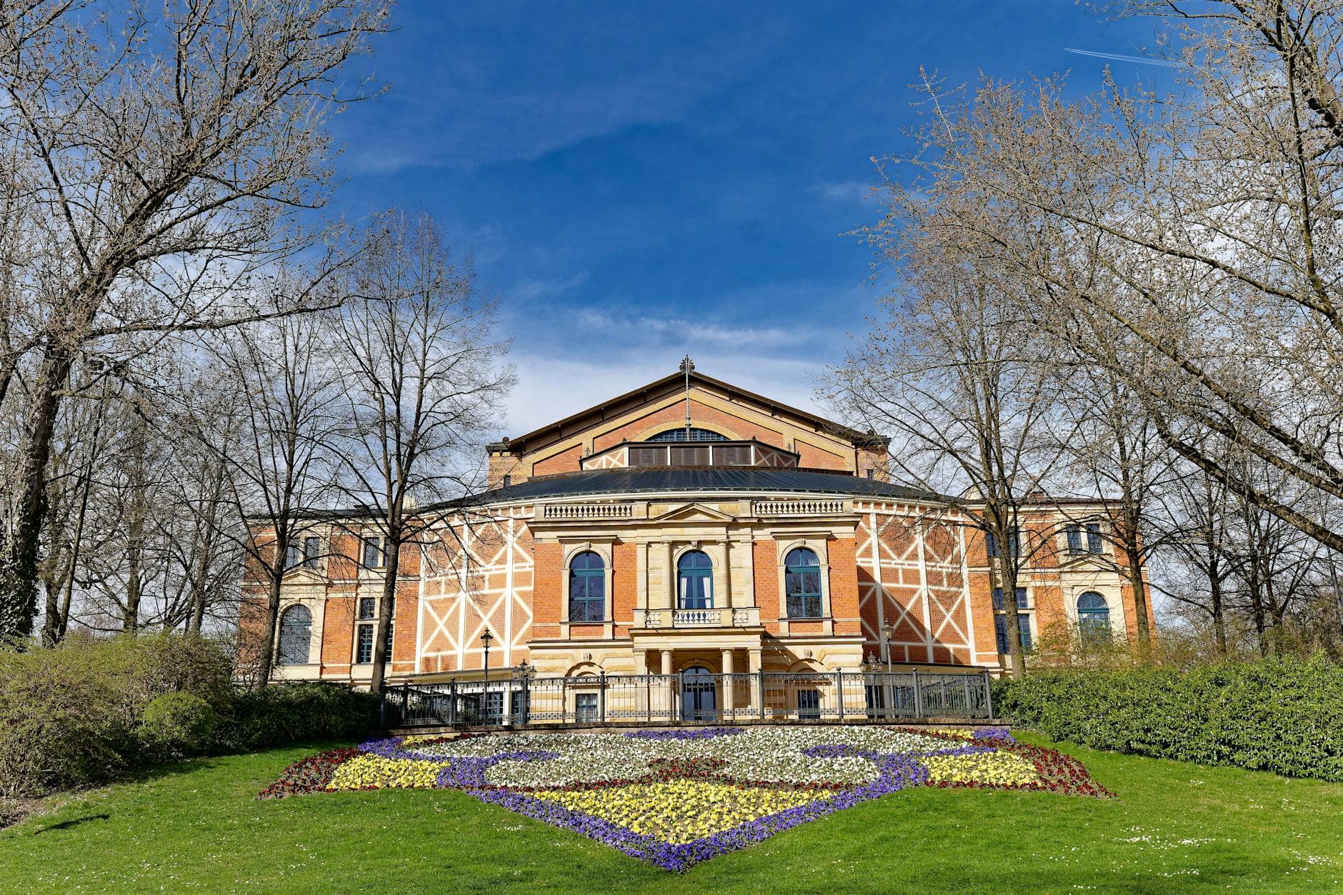 Historisches Backsteingebäude mit Sandsteindetails unter blauem Himmel, umgeben von kahlen Bäumen und einem bunten Blumenbeet auf Wiese.