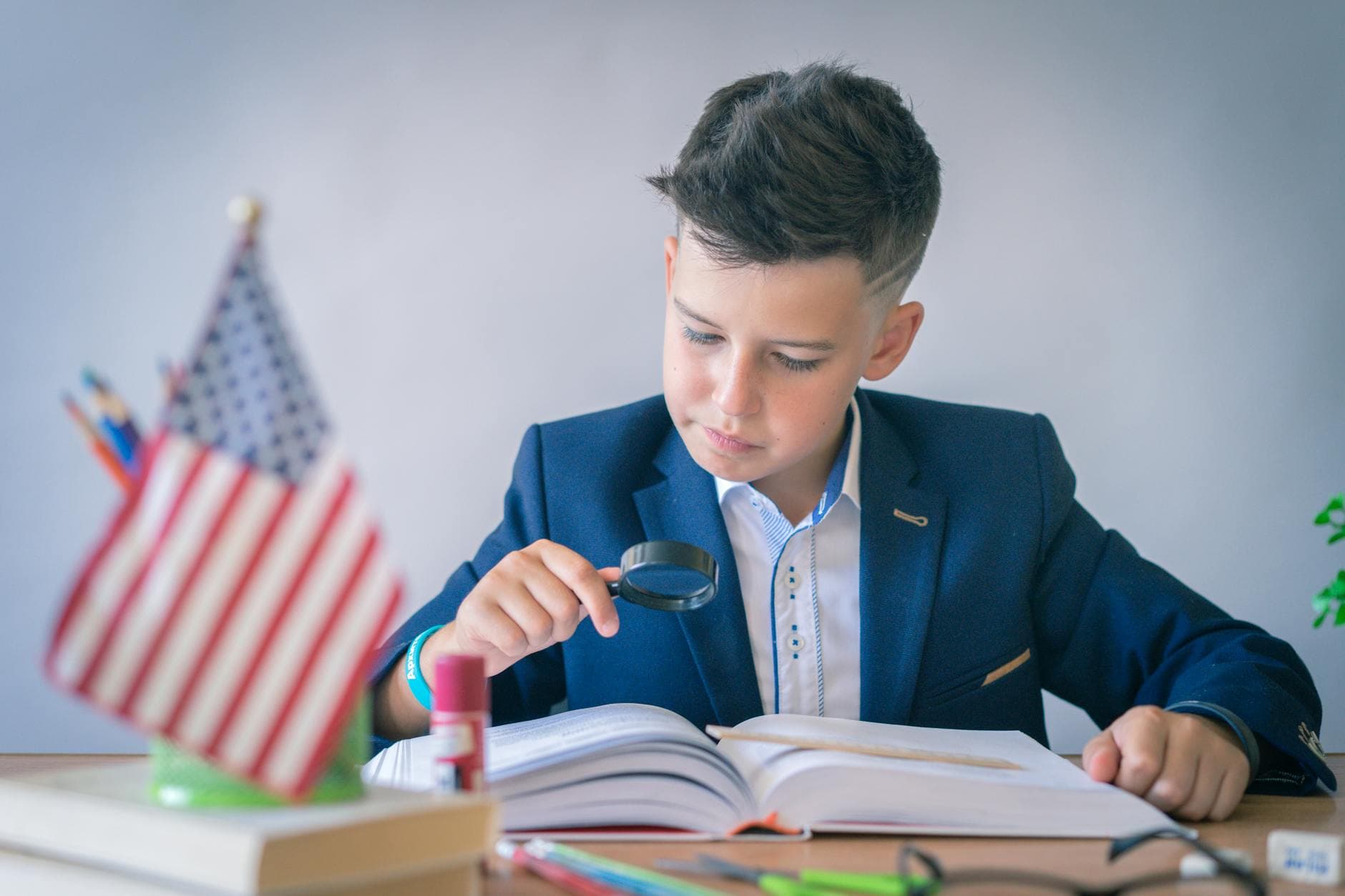 Junge in blauem Anzug liest mit Lupe in Buch am Schreibtisch; daneben kleine US-Flagge, Stifte und Schulmaterial.
