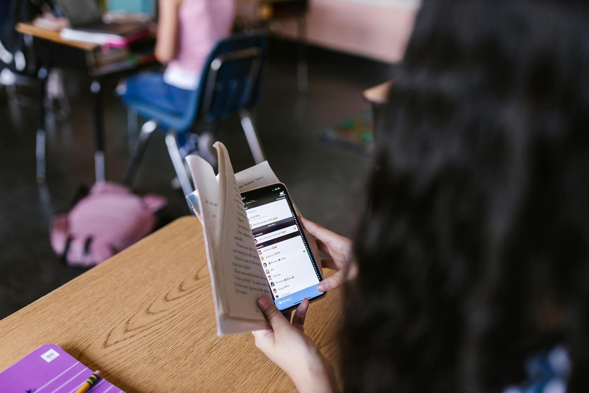 Junge Person hält im Klassenzimmer ein offenes Buch und ein Smartphone mit geöffneter Social-Media-App.
