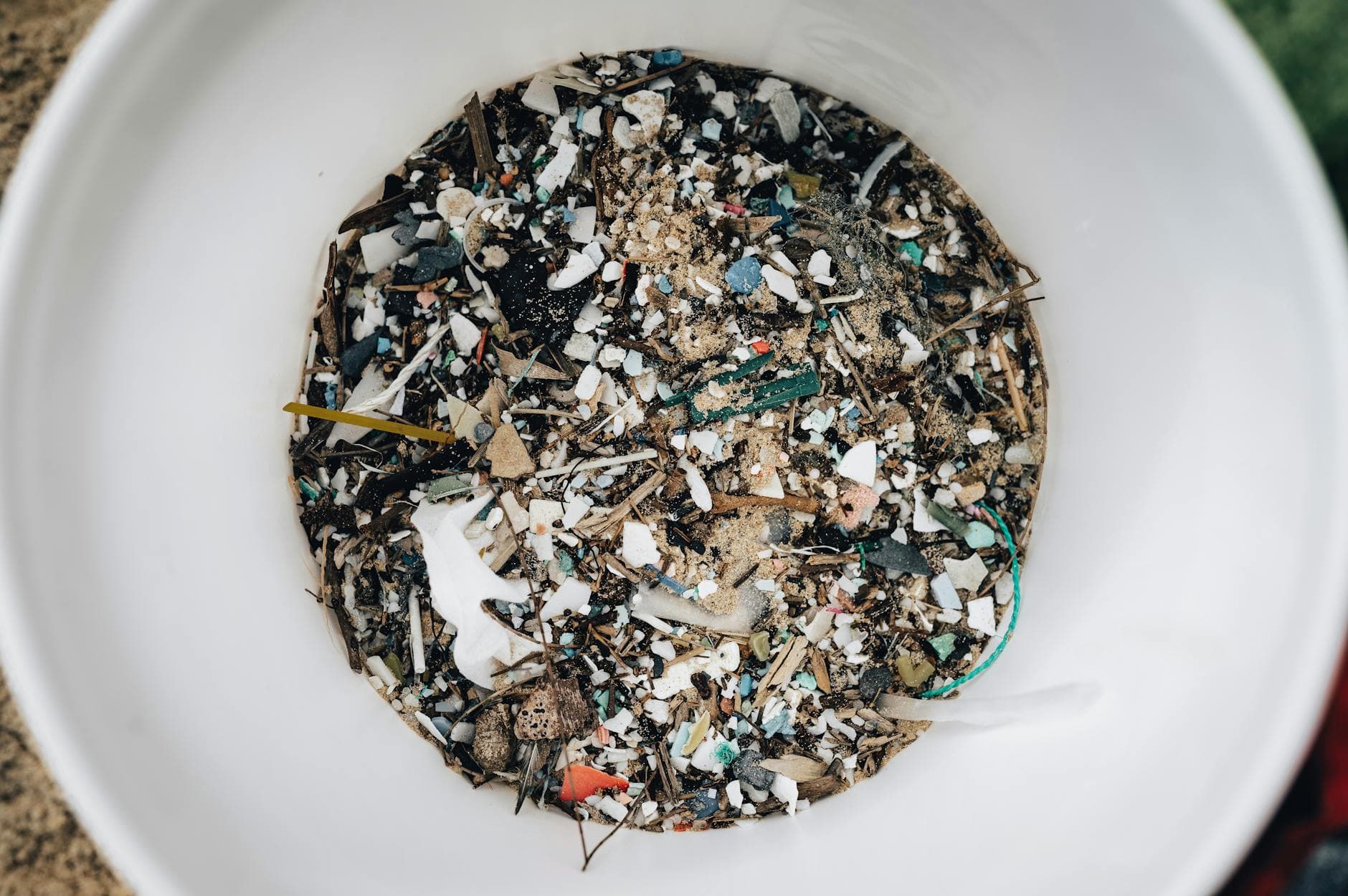 Ein weißer Eimer, von oben gesehen, gefüllt mit Sand, bunten Plastikfragmenten und kleinen Holzstücken.