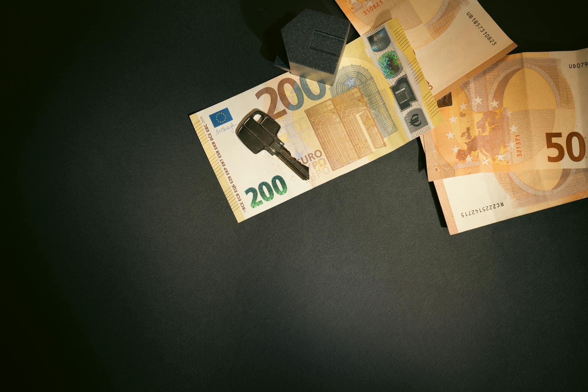 Auf dunklem Untergrund liegen 200- und 50-Euro-Banknoten, darauf ein Schlüssel und dahinter ein schwarzes Haus-Symbol.