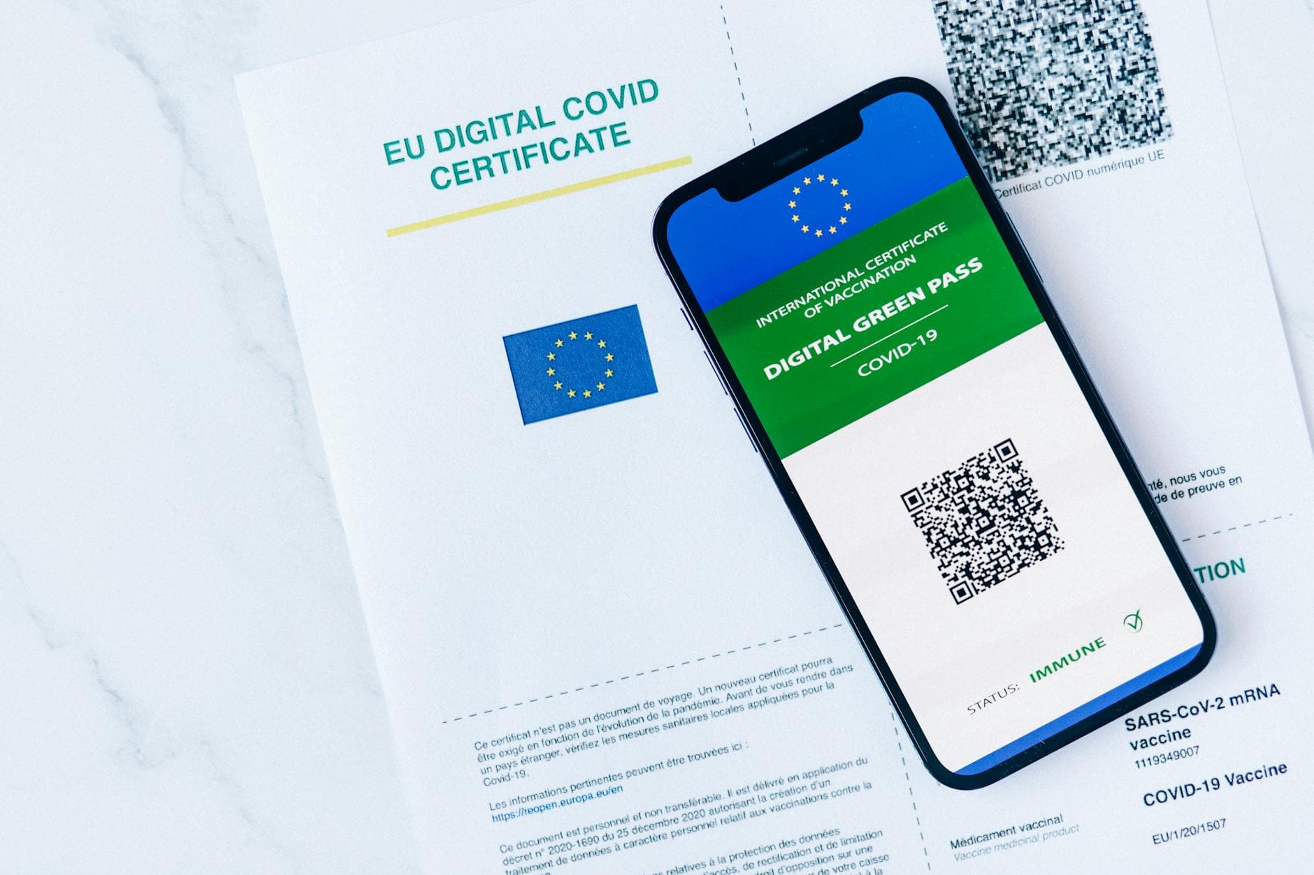 Gedrucktes EU Digitales COVID-Zertifikat neben Smartphone mit EU Digital Green Pass, QR-Code und Status Immune.