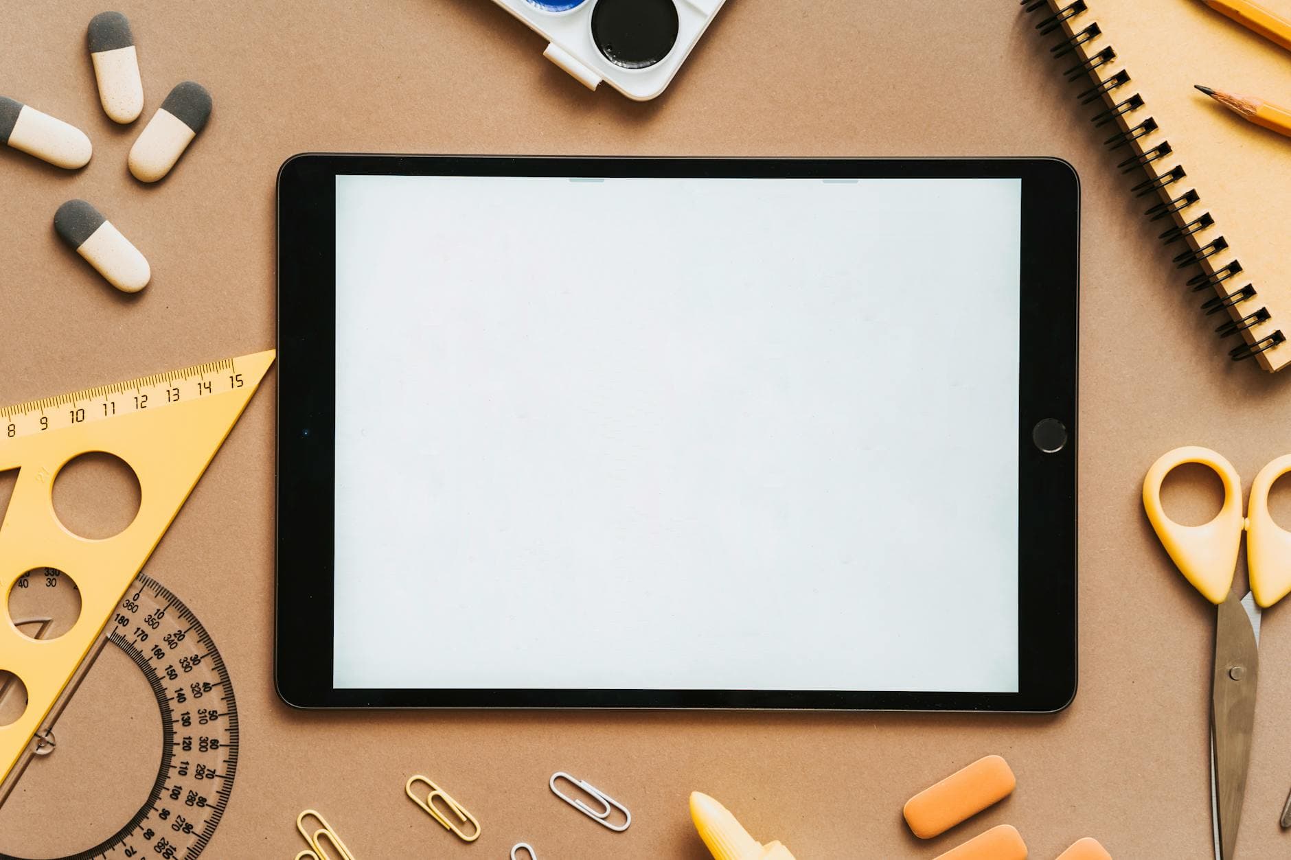 Schwarzes Tablet mit hellem Display, umgeben von diversen Schul- und Büromaterialien auf einem braunen Untergrund.