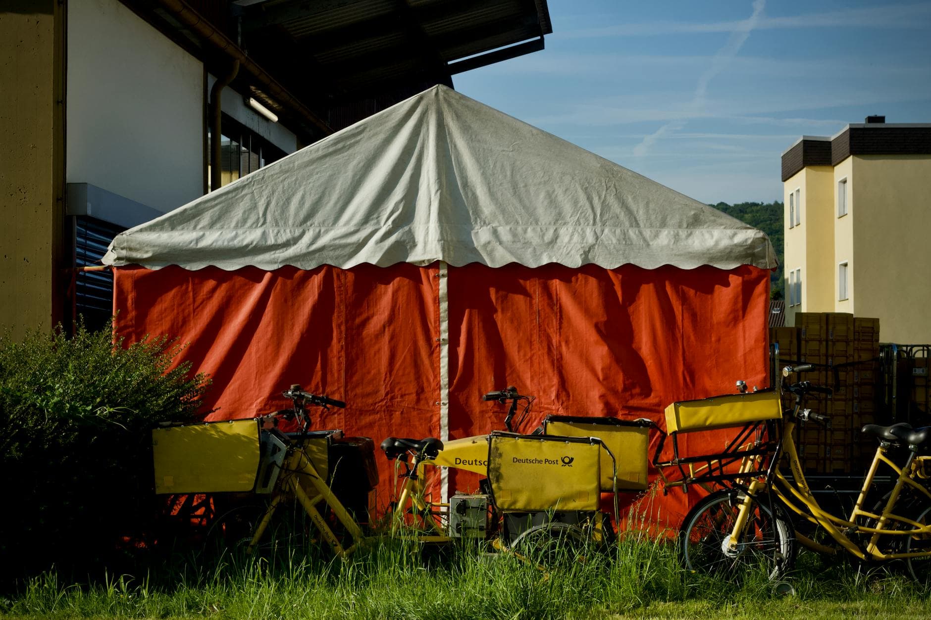 Ein weiß-rotes Zelt, davor gelbe Deutsche Post Fahrräder auf einer Wiese, im Hintergrund Gebäude und blauer Himmel.