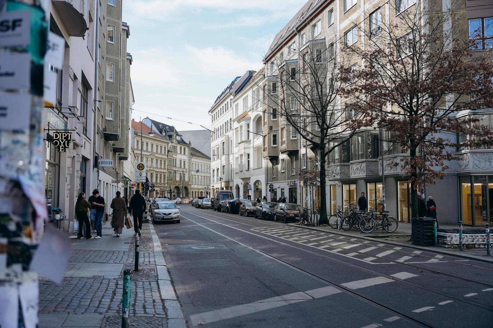 Belebte Stadtstraße mit Altbauhäusern, Straßenbahnschienen, parkenden Autos und Fußgängern am Bürgersteig.