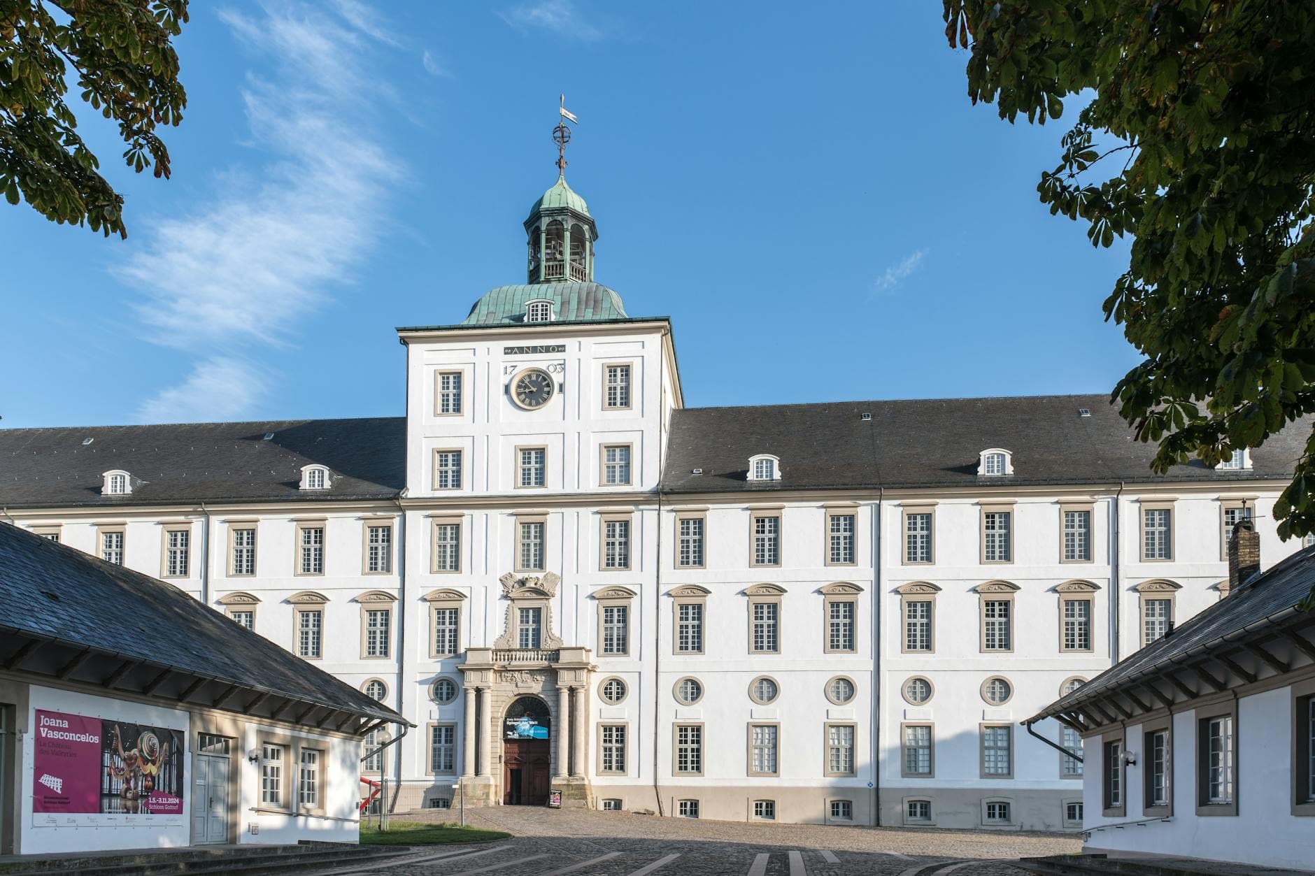 Weißes Schlossgebäude mit Uhrturm, grünem Turmdach, gepflastertem Hof, Nebengebäuden und Bäumen unter blauem Himmel.