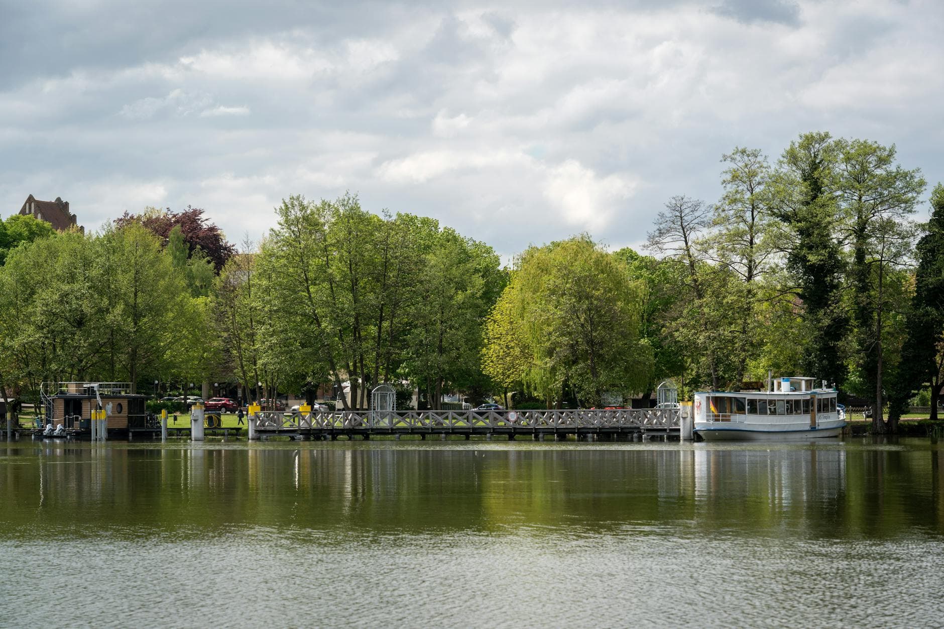 Vom Wasser aus gesehen: bewaldetes Ufer mit langem Holzsteg, einem Hausboot und einem weißen Passagierschiff unter bewölktem Himmel.