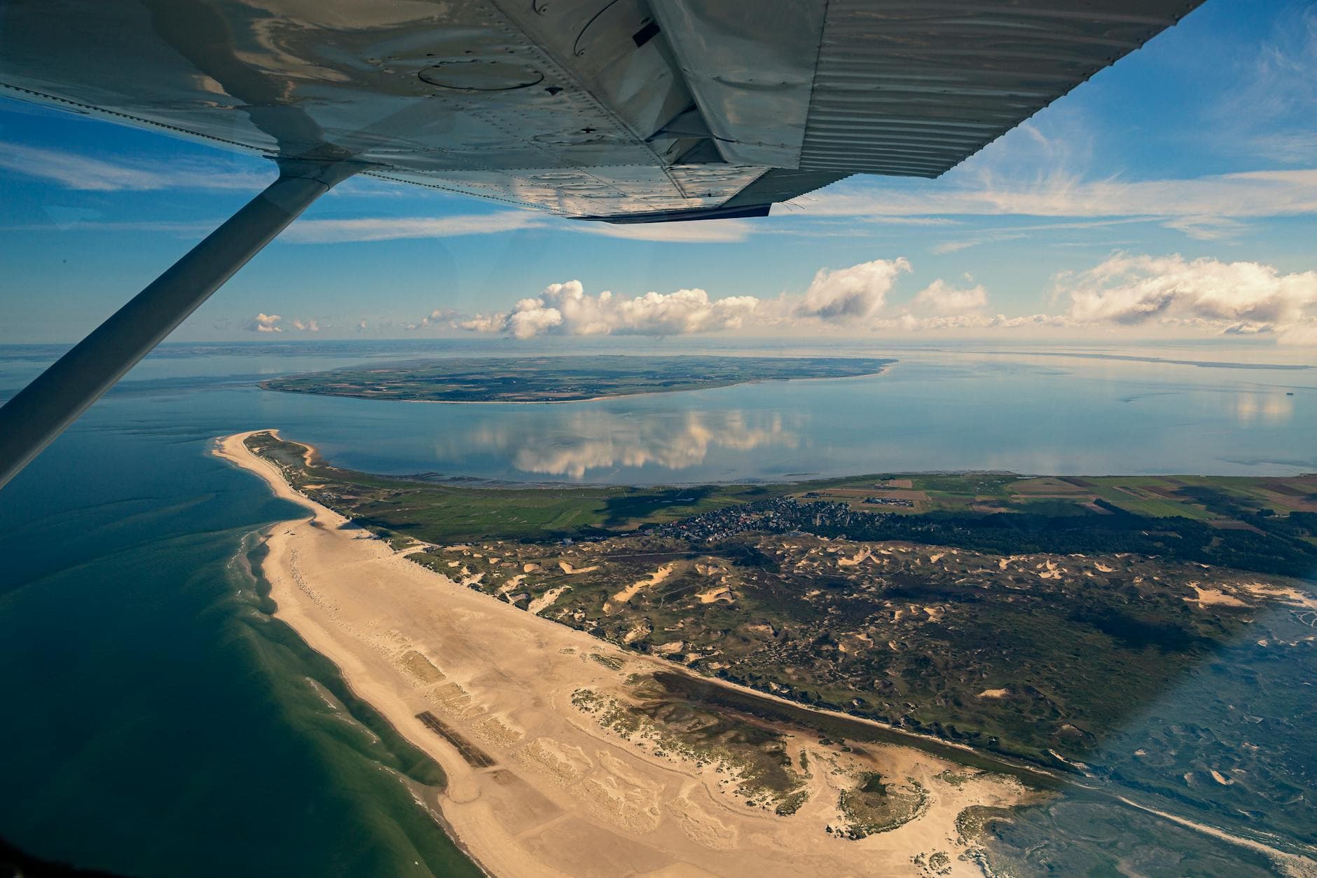 Luftaufnahme von einem Flugzeugflügel aus, mit langem Sandstrand, Dünen, grünem Land, einer Siedlung und dem Meer, das Wolken spiegelt.