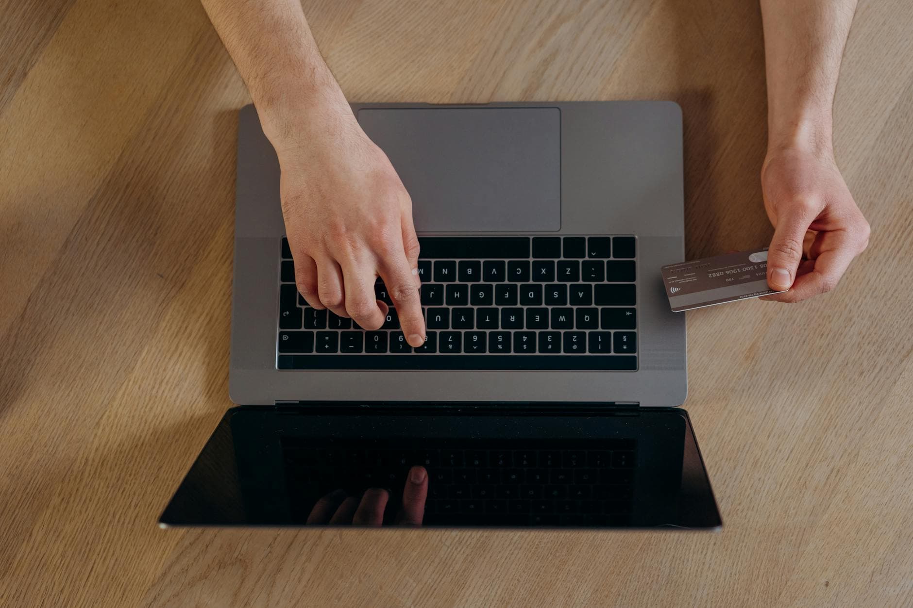Auf einem Holztisch tippt eine Hand auf einem Laptop, die andere hält eine Kreditkarte bereit.