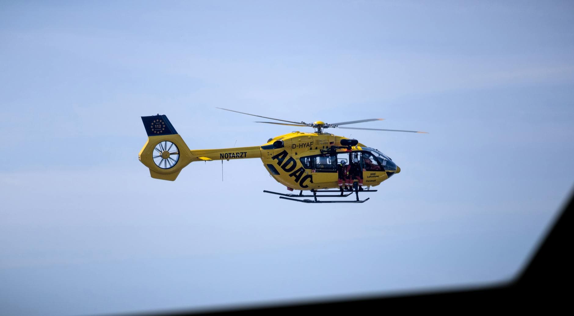 Gelber ADAC-Rettungshubschrauber D-HYAF mit 112-Emblem und Notarzt-Aufschrift fliegt mit offener Tür und Crew vor blauem Himmel.