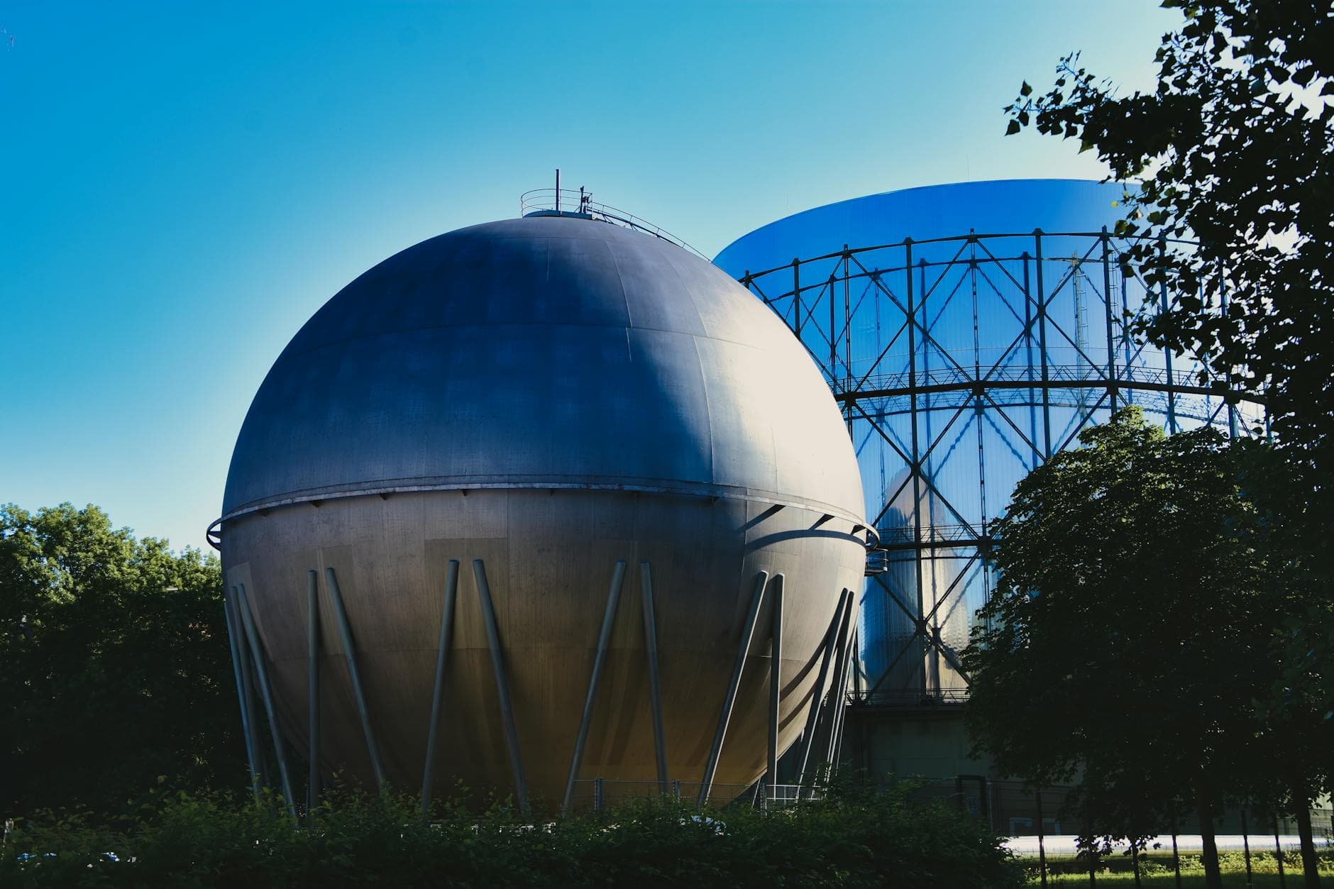 Zwei große Industrietanks, ein grauer Kugeltank und ein blauer mit Gitterstruktur, unter blauem Himmel und Bäumen.