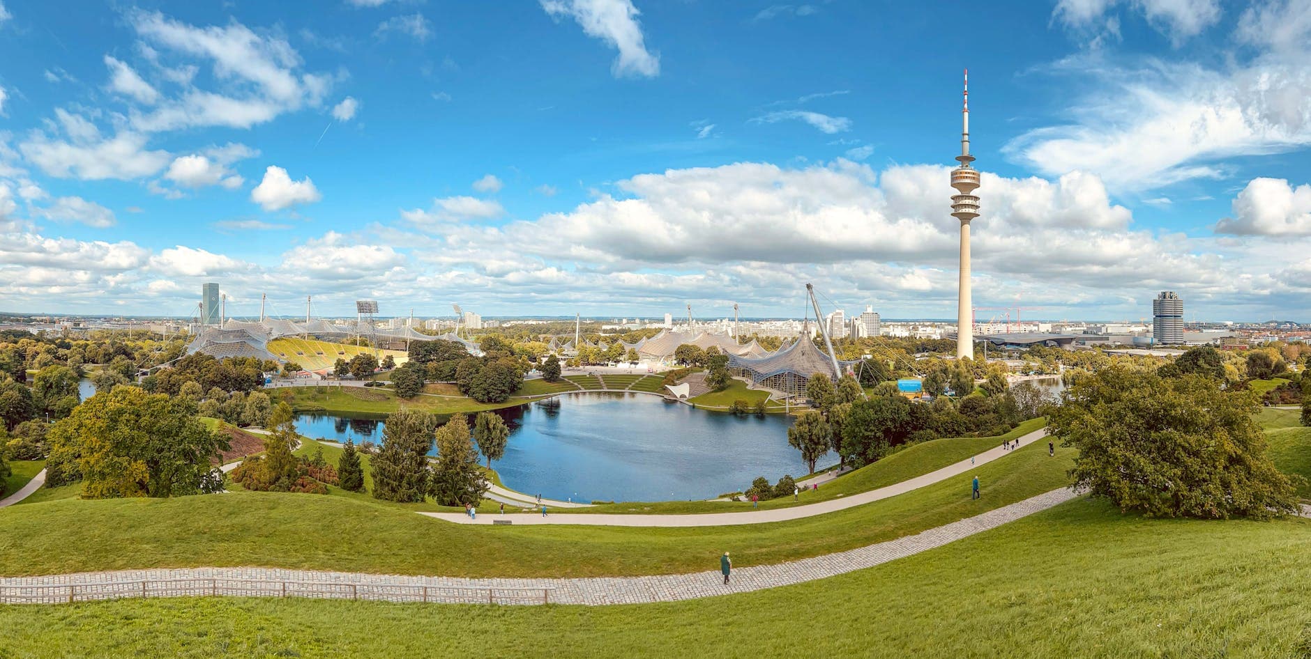 Olympiapark München mit Olympiastadion, Olympiasee, Olympiaturm, grünen Hügeln, Spaziergängern und Stadtpanorama unter blauem Himmel.