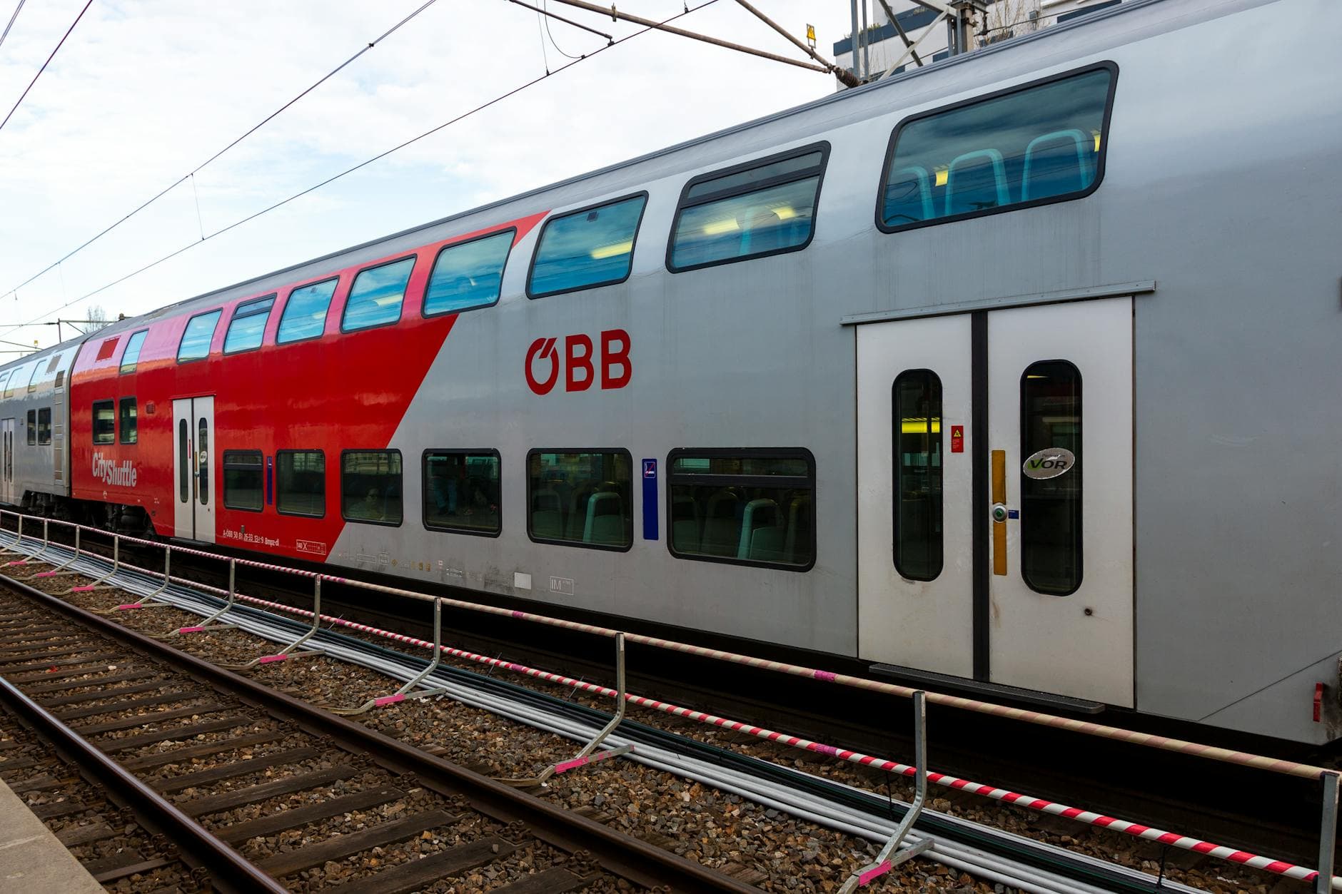 Ein roter und silberner ÖBB-Doppelstockzug steht auf Bahngleisen, mit Oberleitungen und Absperrgittern.