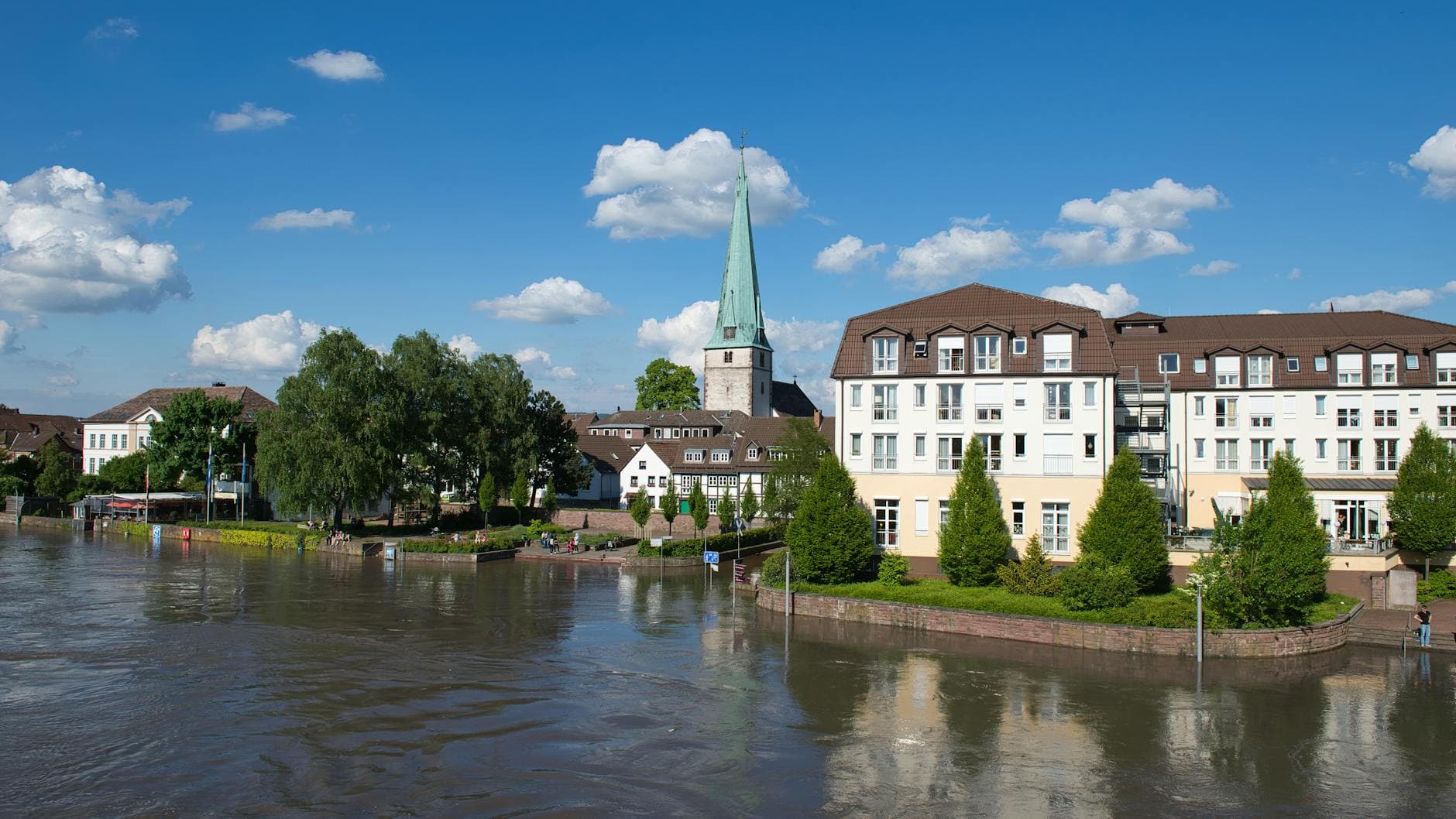Panoramablick auf eine Stadt am Fluss mit schmutzigem Wasser, einer Kirche mit grünem Turm, alten und neuen Häusern, Menschen und Wolken.