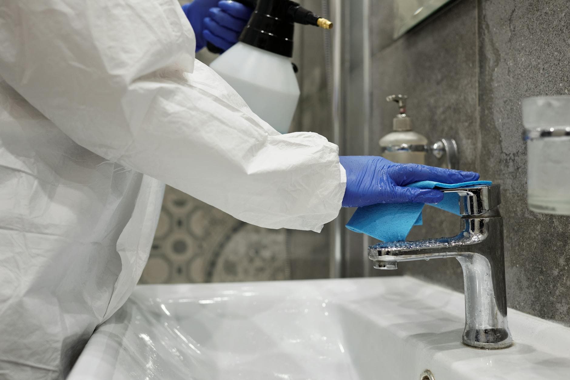 Person in weißem Schutzanzug und blauen Handschuhen säubert Wasserhahn mit blauem Lappen; hält Sprühflasche.