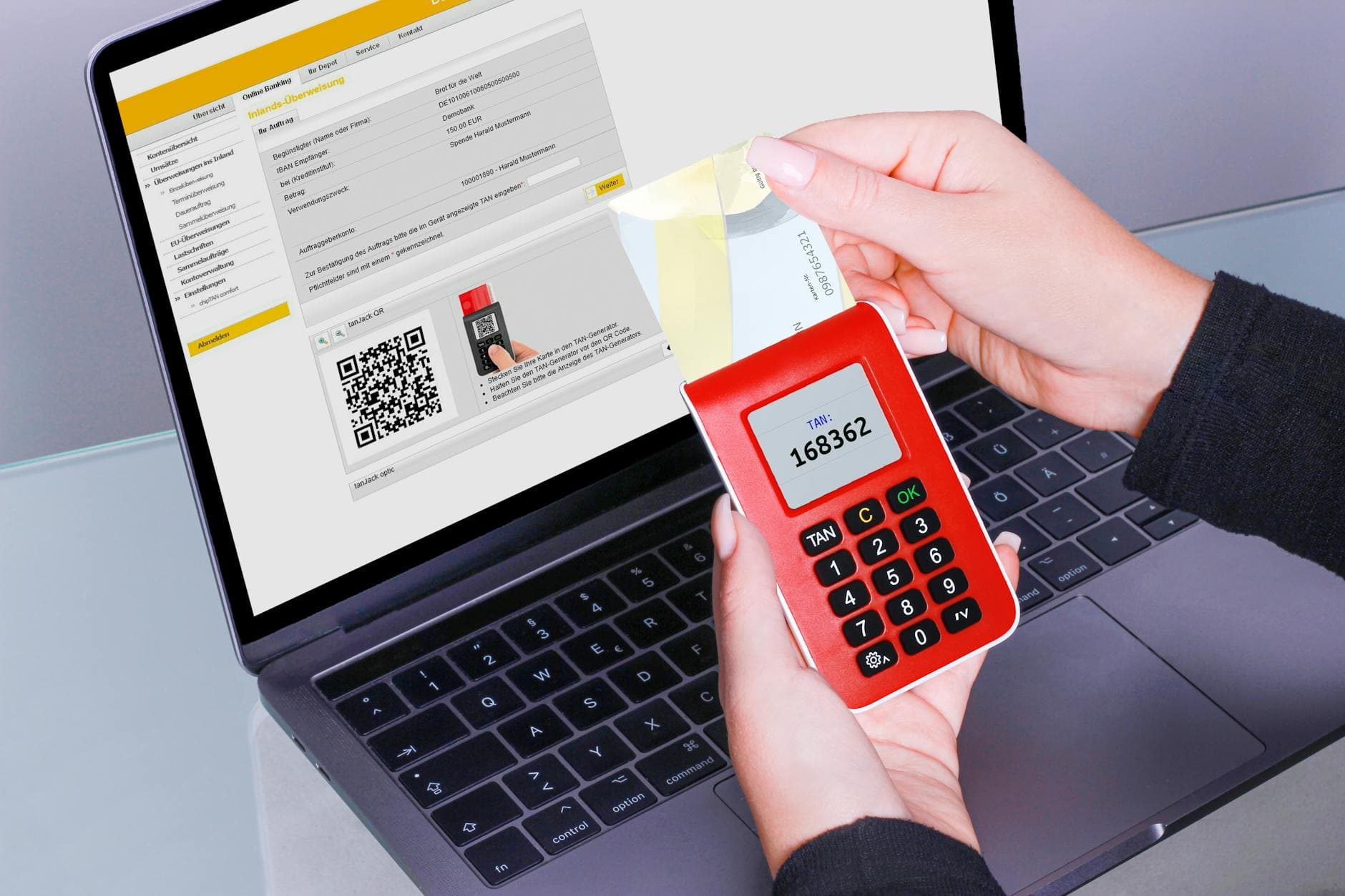 Eine Person hält einen roten TAN-Generator mit einer Bankkarte vor einem Laptop, dessen Bildschirm eine Online-Banking-Überweisung zeigt.